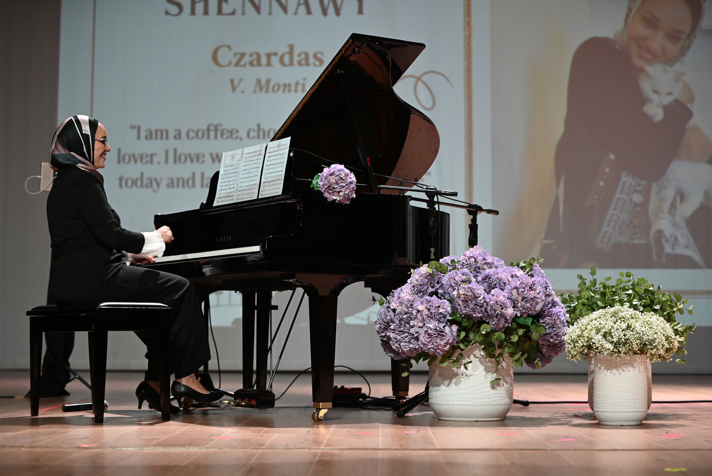 Yasmin Shennawy playing a grand piano on stage beside hydrangeas